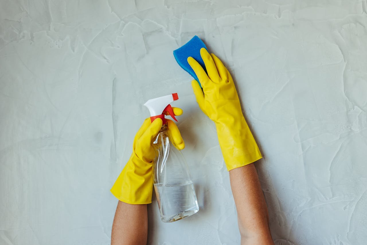 Close-up of hands in yellow gloves cleaning a wall with a spray bottle and sponge.